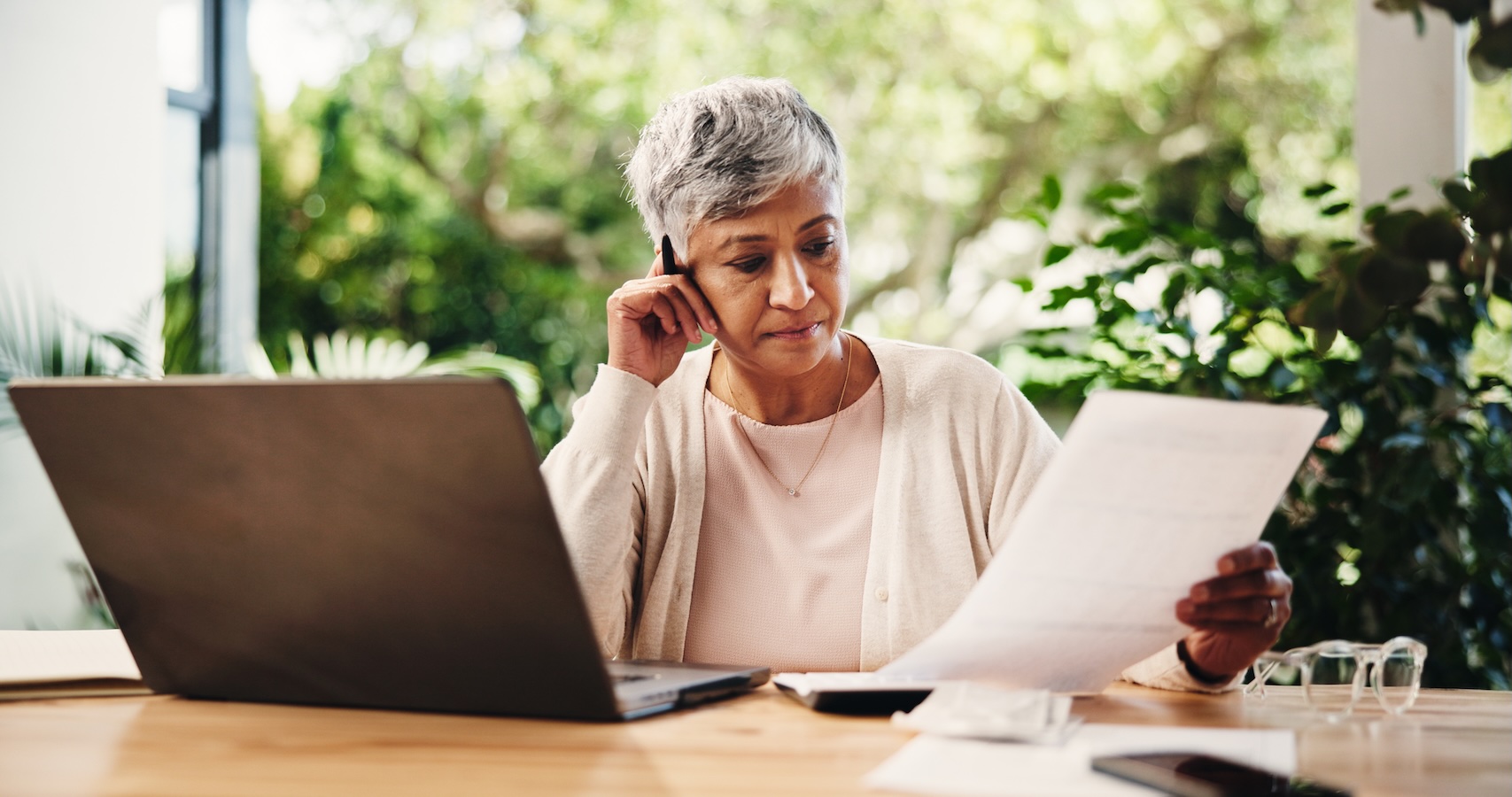 Senior woman at home sorting through documents while using laptop and looking serious.